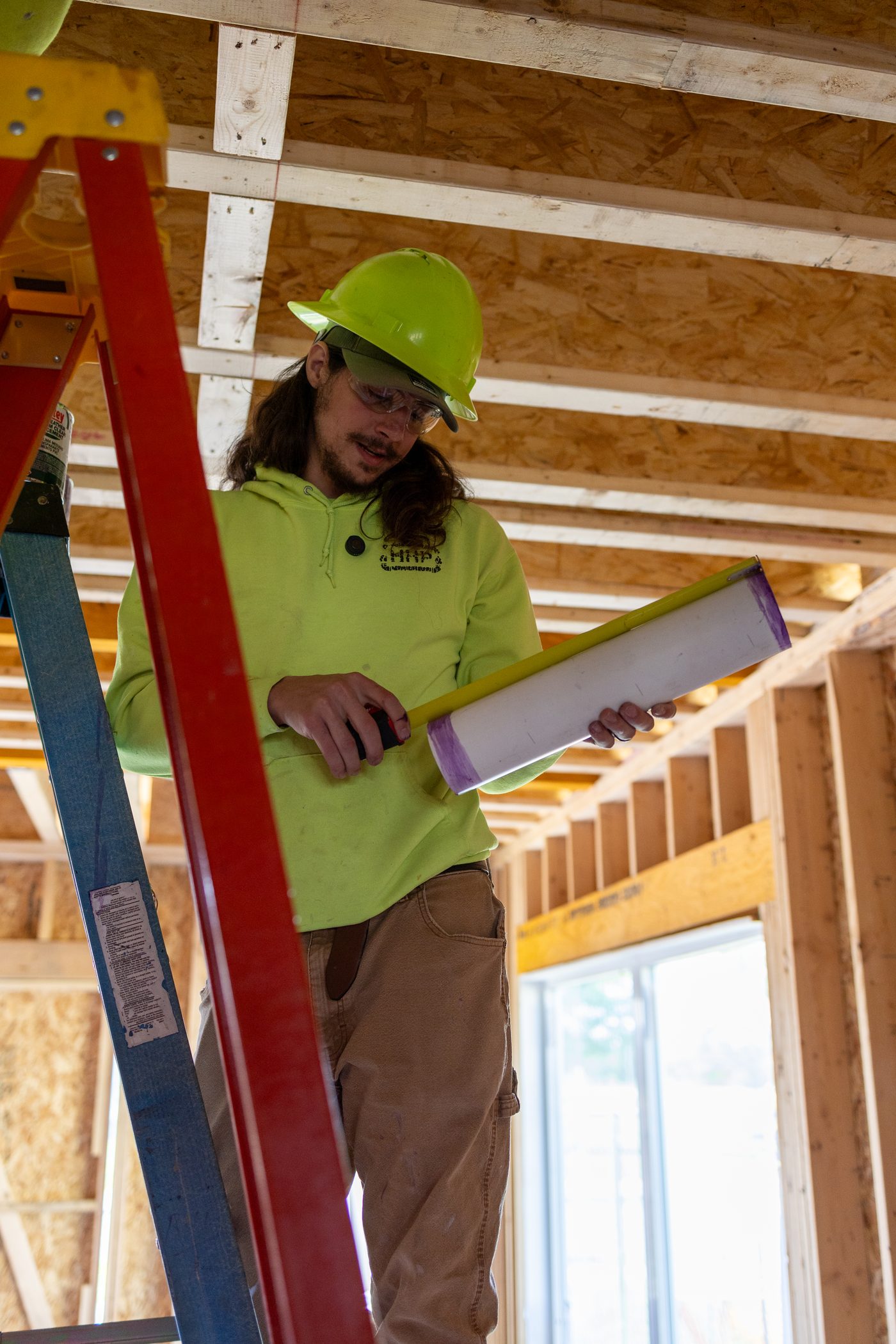 A Hertel plumber in a hard hat priming PVC pipe inside a new-build frame